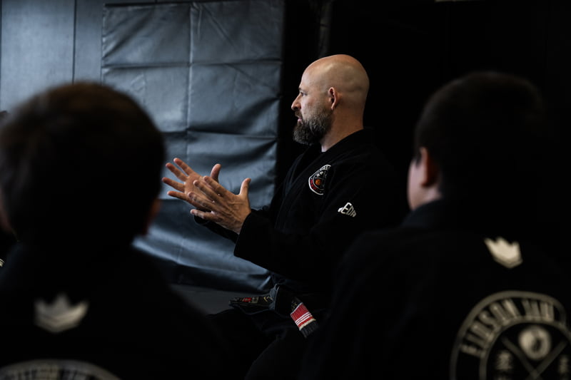 Instructor teaching martial arts class – Jiu Jitsu San Diego, CA A martial arts instructor in a black gi explains a Jiu Jitsu technique to students seated attentively on the mat during class – Jiu Jitsu San Diego, CA
