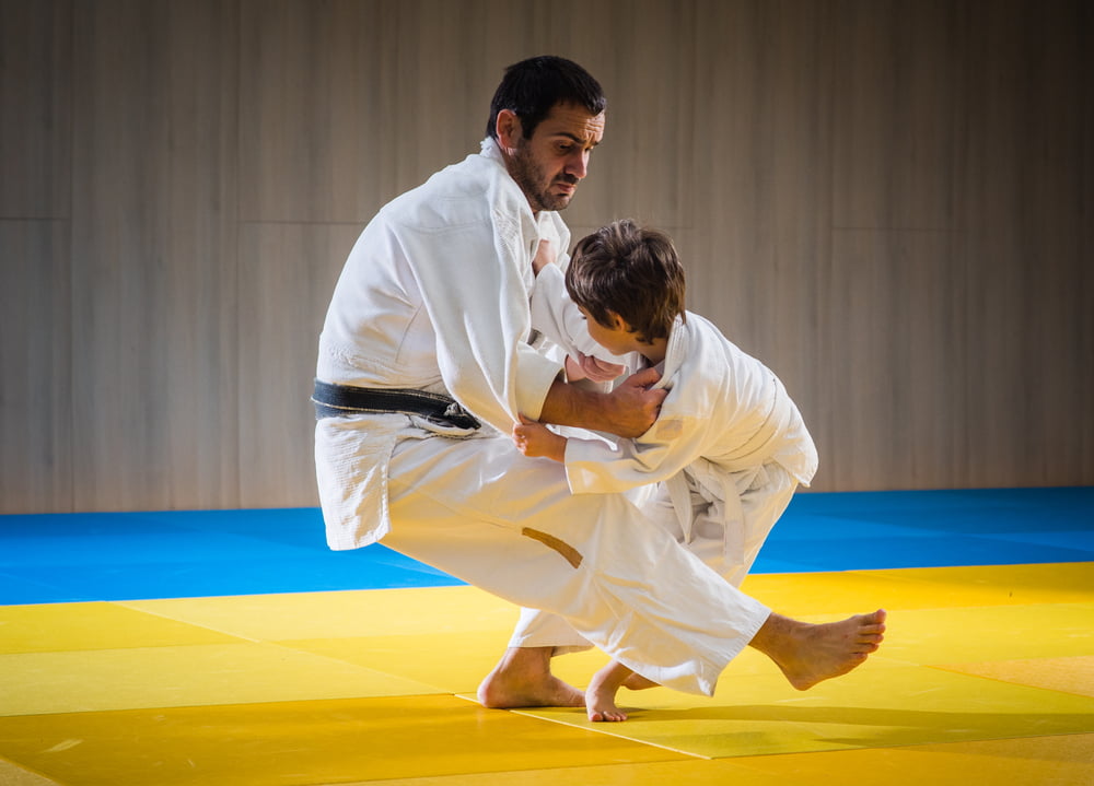 Adult martial arts instructor practicing a self defense takedown move with young student on yellow mat - Self Defense
